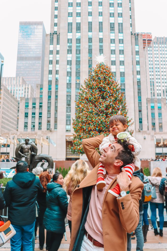 Man with a child on his shoulders standing in front of the Christmas tree at Rockefeller center. 