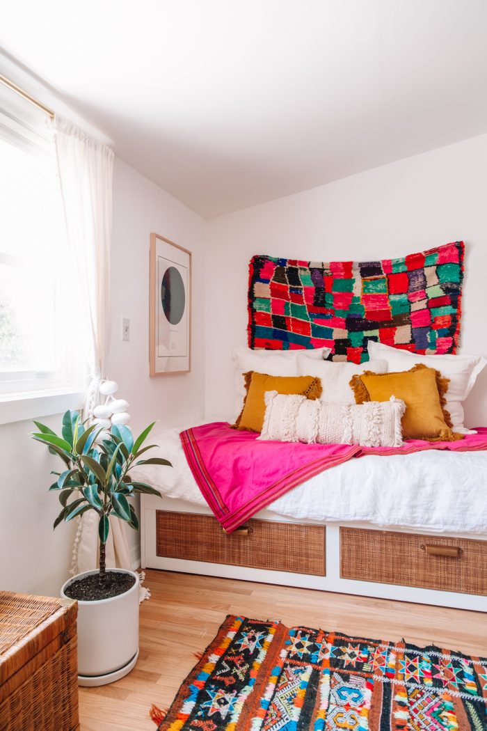 IKEA bed with rattan cane on the front and white bedding with a pink blanket and a plant in front of it