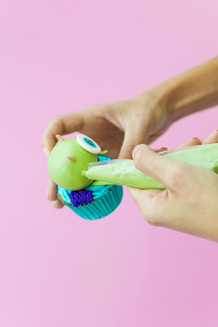 Green Mike Wazowski cake ball onto of a cupcake with icing being placed on