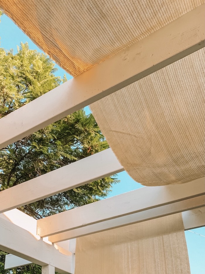 Pergola sun shade hanging on white beams with green tree in background