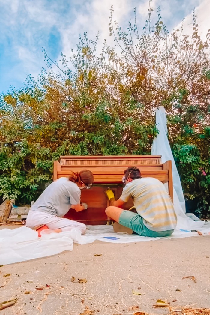 Man and woman sanding a wood piano outside in front of greenery