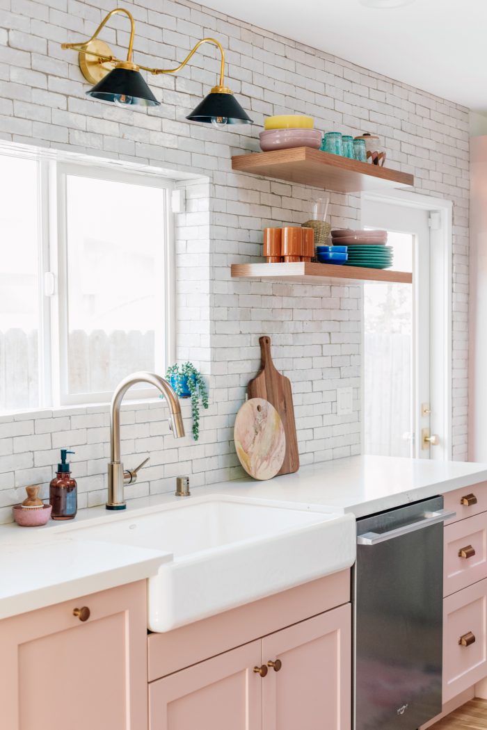 White tile kitchen with light pink cabinets and colorful glassware on floating shelves