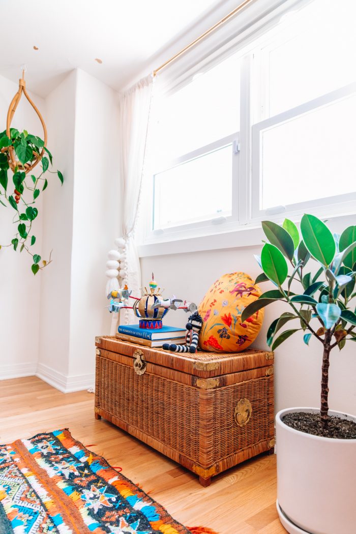 wicker trunk with pillow and books on top under a window next to two plants and a colorful moroccan rug